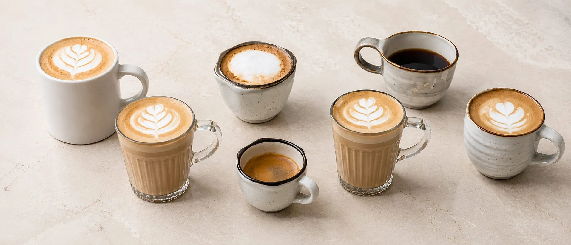 Tazas de café de especialidad sobre una mesa de mármol: varias con arte en leche, un espresso corto y un americano