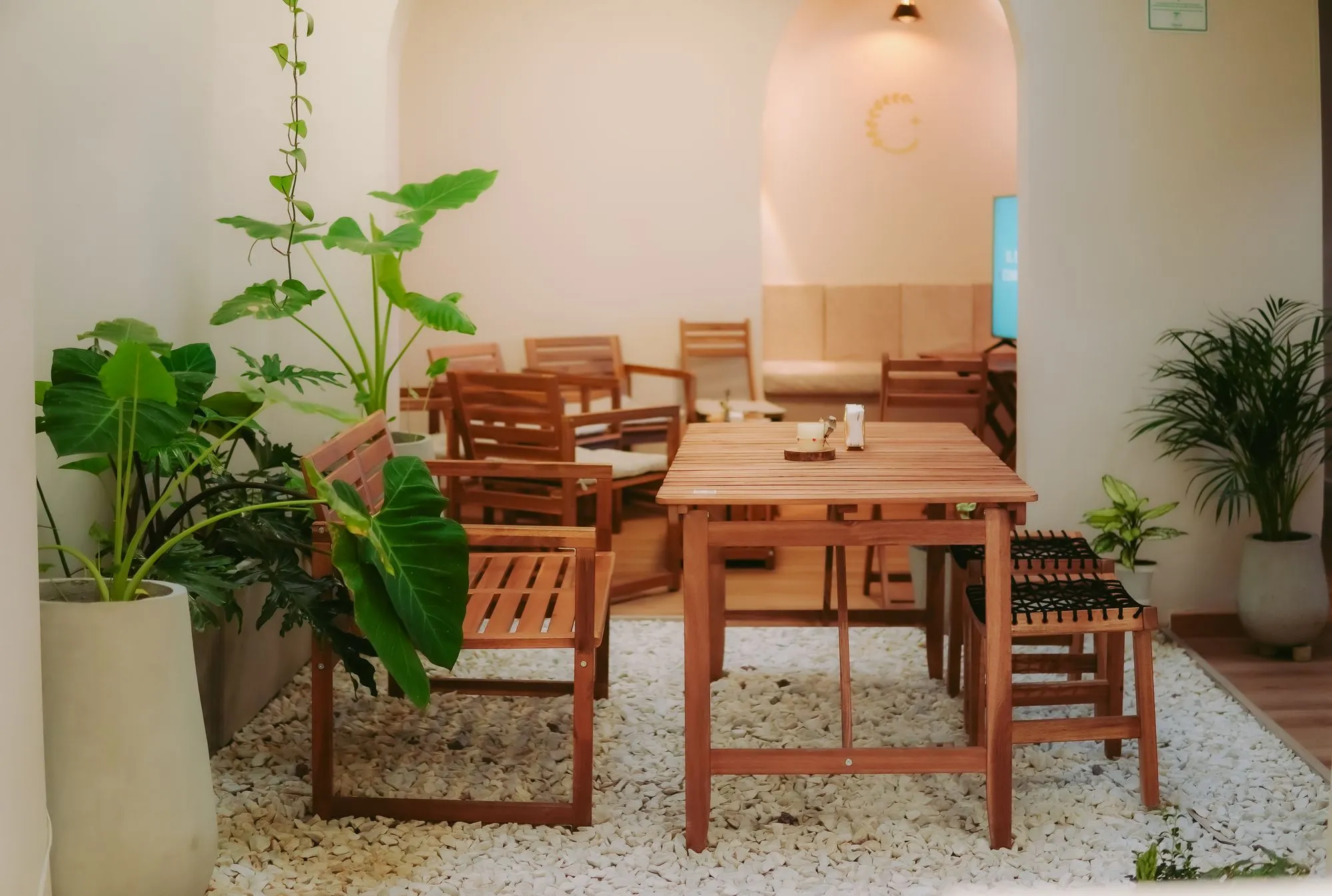 Mesas de madera sobre gravilla entre plantas grandes, con un arco iluminado al fondo, en el patio interno de CAELUS